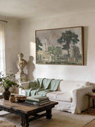 Framed vintage European landscape art mounted above a white linen sofa with a sage green throw in a sunlit living room.